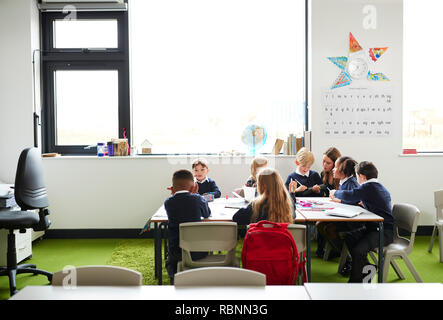 A group of primary school kids sitting at table in a classroom, teacher kneeling to help them during the lesson Stock Photo