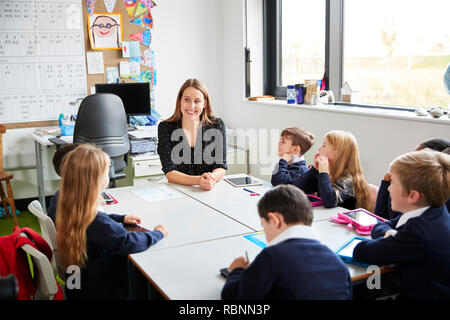 Primary school children sitting at a table in a classroom with their female teacher, looking at each other