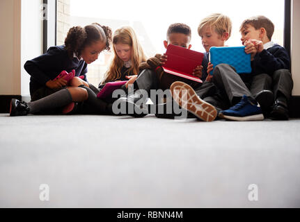Low angle view of primary school friends sitting together in front of a window in a school corridor looking at tablet computers together Stock Photo