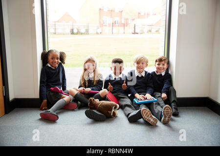 Five primary school friends with tablet computers sitting on the floor in a school corridor at break time, smiling to camera, selective focus Stock Photo