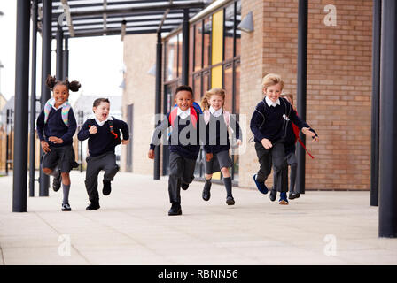 Primary school kids, wearing school uniforms and backpacks, running on a walkway outside their school building, front view Stock Photo