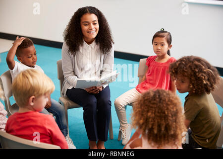 Young female school teacher reading a book to kindergarten children, sitting on chairs in a circle in the classroom listening, close up Stock Photo