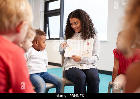 Young female teacher showing a picture in a book to kindergarten children sitting on chairs in a circle in the classroom, close up, selective focus Stock Photo