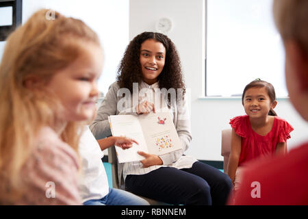 Over shoulder view of female teacher showing a picture in a book to a group of kindergarten children sitting on chairs in a classroom, close up Stock Photo