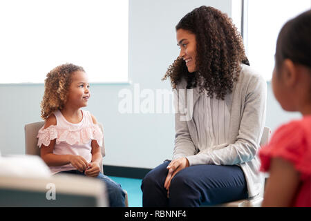 Infant school girl and her female teacher sitting on chairs in a classroom smiling to each other, close up