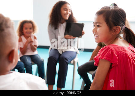 Over shoulder view of smiling young female school teacher showing a tablet computer to infant school children, sitting in a circle in the classroom, over shoulder view Stock Photo