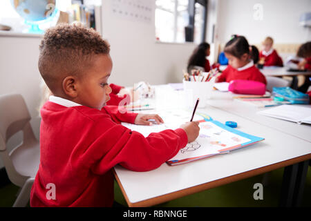 Young black schoolboy wearing school uniform sitting at a desk in an infant school classroom drawing, close up, side view Stock Photo