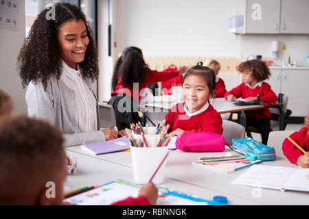 Female teacher and Chinese schoolgirl sitting at a table in an infant school class smiling to other kids, selective focus Stock Photo