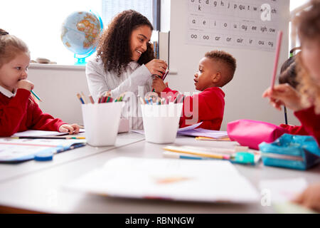 Smiling millennial female infant school teacher sitting at table with kids in a classroom giving pencils to a schoolboy, low angle Stock Photo