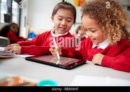 Close up of two kindergarten schoolgirls wearing school uniforms, sitting at a desk in a classroom using a tablet computer and stylus, looking at screen and smiling Stock Photo