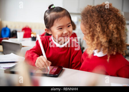 Close up of two kindergarten schoolgirls wearing school uniforms, sitting at a desk in a classroom using a tablet computer and stylus, looking at each other smiling Stock Photo