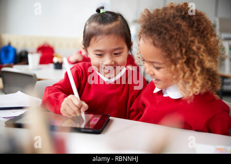 Close up of two kindergarten schoolgirls wearing school uniforms, sitting at a desk in a classroom using a tablet computer and stylus, looking at the screen and smiling Stock Photo