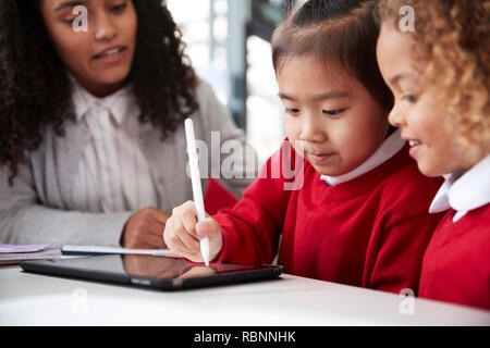 Close up of female infant school teacher sitting at a desk in a classroom helping two schoolgirls wearing school uniforms using a tablet computer and stylus, looking at screen and smiling Stock Photo