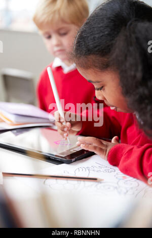 Close up of a schoolgirl sitting at desk next to a boy in an infant school classroom, using a tablet computer and stylus, selective focus, vertical Stock Photo