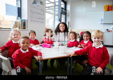 Group portrait of infant school teacher and kids sitting at table in a classroom looking to camera smiling, front view Stock Photo