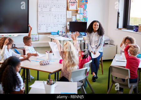 Young female infant school teacher sitting on a chair facing school kids in a classroom, raising their hands to answer a question Stock Photo