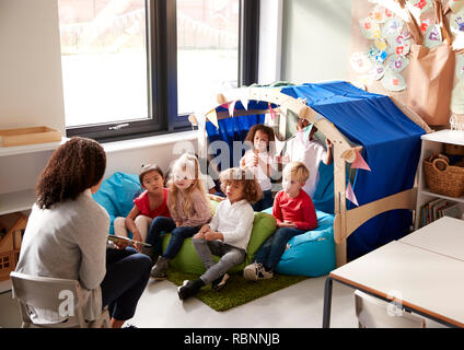 Female infant school teacher sitting on a chair showing a book to a group of children sitting on bean bags in a comfortable corner of the classroom, elevated view Stock Photo