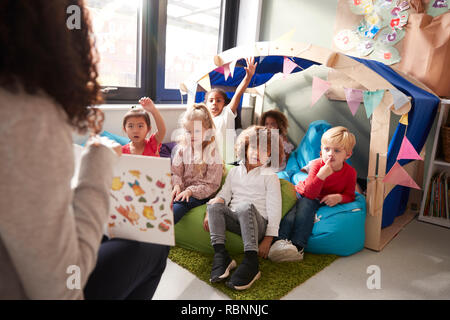 Female infant school teacher sitting on a chair showing a book to a group of children sitting on bean bags in a comfortable corner of the classroom, raising their hands to answer a question, elevated view, close up Stock Photo