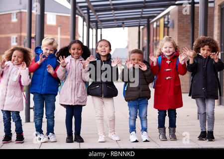 A group of smiling young multi-ethnic school kids wearing coats and carrying schoolbags standing in a row in walkway outside their infant school waving to camera, full length, front view Stock Photo