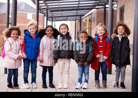 A group of smiling young multi-ethnic school kids wearing coats and carrying schoolbags standing in a row in walkway outside their infant school smiling to camera, full length, front view Stock Photo