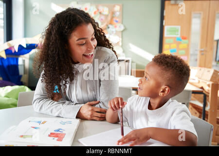 Female infant school teacher working one on one with a young schoolboy, sitting at a table smiling at each other, close up Stock Photo