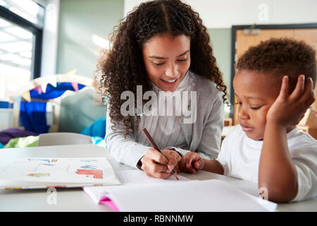 Female infant school teacher working one on one with a young schoolboy, sitting at a table writing with him, close up Stock Photo