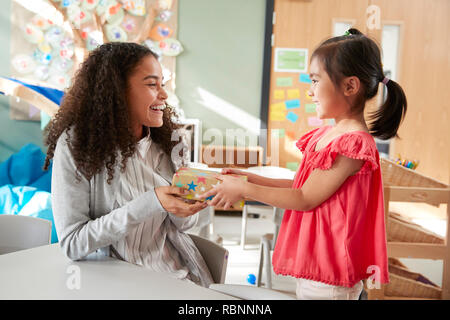 Kindergarten schoolgirl giving a gift to her female teacher in a classroom, side view, close up Stock Photo
