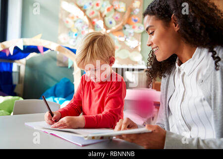 Female infant school teacher working one on one with a young white schoolboy, sitting at a table in a classroom writing, close up Stock Photo