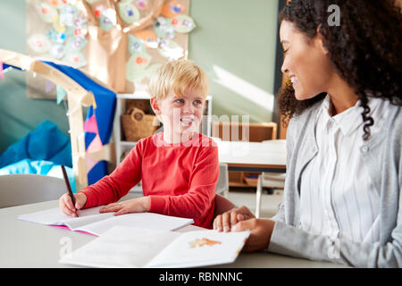 Female infant school teacher working one on one with a young white schoolboy, sitting at a table in a classroom writing, looking at each other, close up Stock Photo