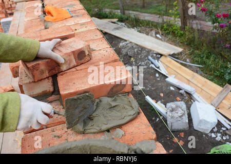 Bricklaying. Bricklayer worker installing red blocks and caulking brick masonry joints exterior brick house  wall with trowel putty knife outdoor.  Ba Stock Photo