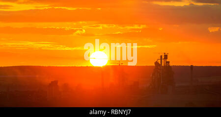 Dark silhouettes of tall buildings of modern cement plant and power station on bright big white setting or rising sun in dramatic yellow red sky backg Stock Photo