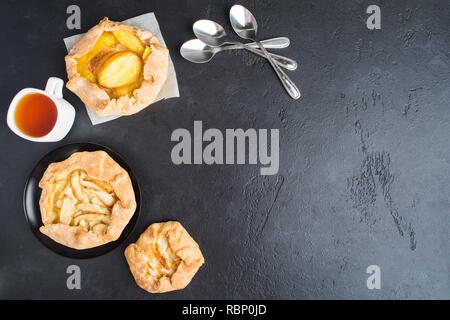 Homemade sweet baking with apples and appricots with one cup of tea and spoons over black background with copy space Stock Photo