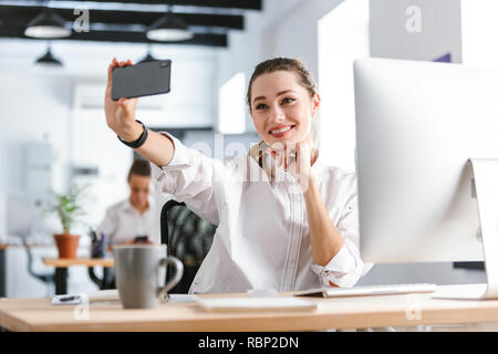 Shocked young businesswoman dressed in shirt sitting at her workplace at the office, taking a selfie Stock Photo
