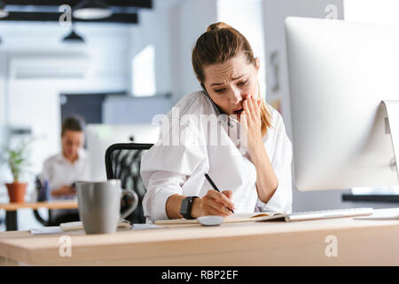 Shocked young businesswoman sitting at her workplace at the office, talking on mobile phone, taking notes Stock Photo