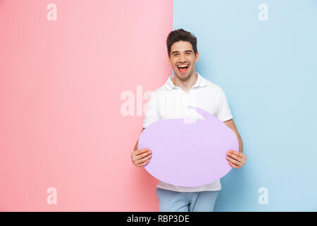Excited young man standing isolated over two colored background, holding empty speech bubble Stock Photo