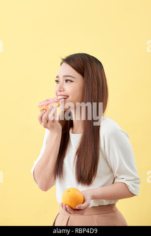 Dieting concept. Beautiful Young Asian Woman holding orange and donut over yellow background. Choosing between junk food with healthy food. Stock Photo