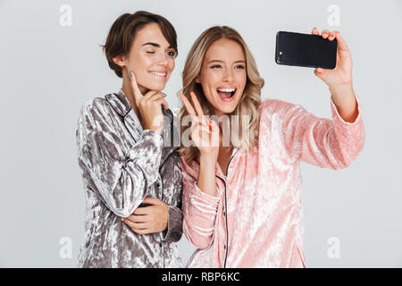 Two pretty girls wearing pajamas isolated over gray background, taking a selfie Stock Photo