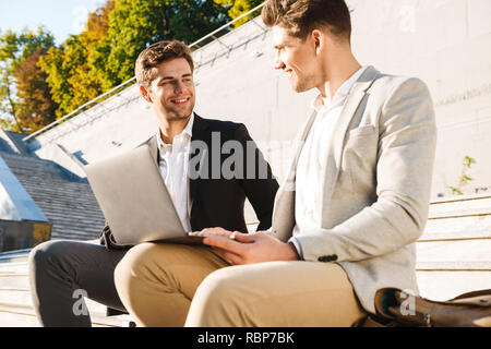 Two confident businessmen using laptop computer while sitting on a bench outdoors Stock Photo