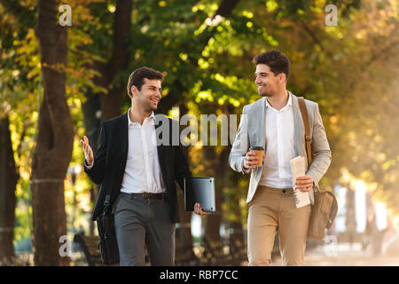 Two happy businessmen walking outdoors, drinking coffee, talking Stock Photo