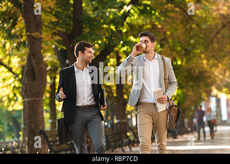 Two happy businessmen walking outdoors, drinking coffee, talking Stock Photo