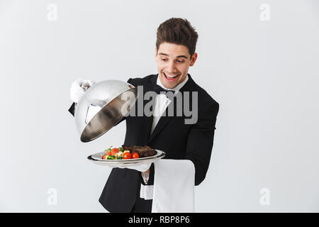 Portrait of a handsome young waiter in tuxedo showing beef steak dish on a plate over white background Stock Photo