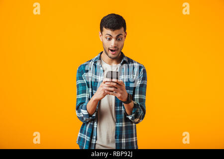 Surprised young man wearing plaid shirt standing isolated over orange background, using mobile phone Stock Photo