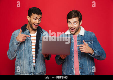 Two cheerful men friends standing isolated over red background, holding laptop computer, celebrating Stock Photo