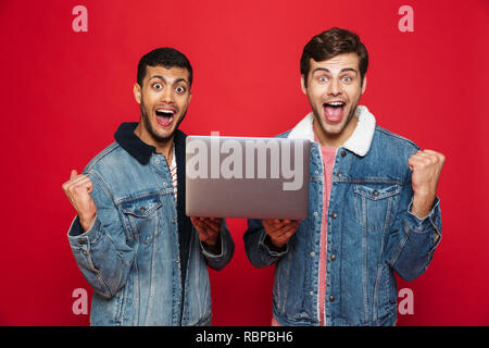 Two cheerful men friends standing isolated over red background, holding laptop computer, celebrating Stock Photo