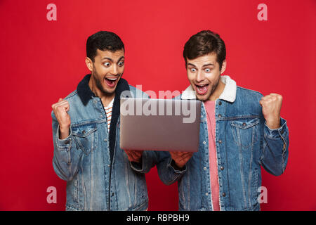 Two cheerful men friends standing isolated over red background, holding laptop computer, celebrating Stock Photo