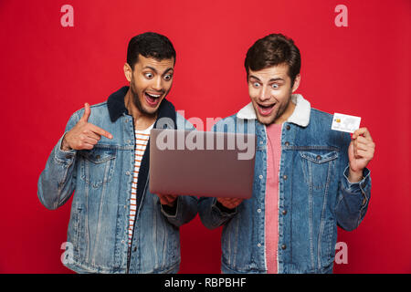 Two cheerful men friends standing isolated over red background, holding laptop computer, showing plastic credit card Stock Photo