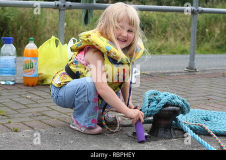 happy blonde little girl, kid wearing lifejacket life vesting, water safety concept rope, ropes, dock, boat moored, sail, sailing, picnic sailing Stock Photo