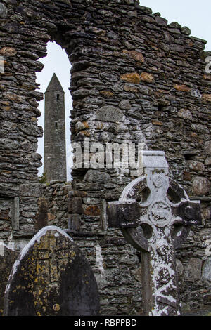 The Round Tower viewed through a window in a stone wall with a Celtic cross and grave stones in the foreground at the Glendalough Monastic Site in Wic Stock Photo