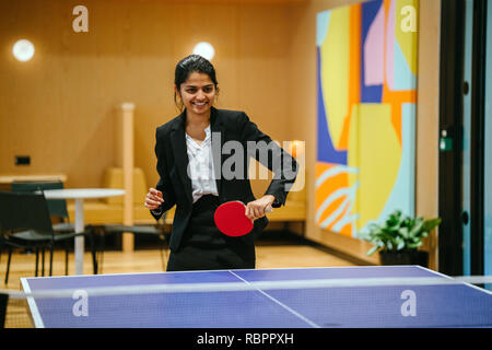 A young Indian Asian woman in a suit smiles and laughs as she plays ping pong in her startup office. She is having lots of fun with a colleague. Stock Photo