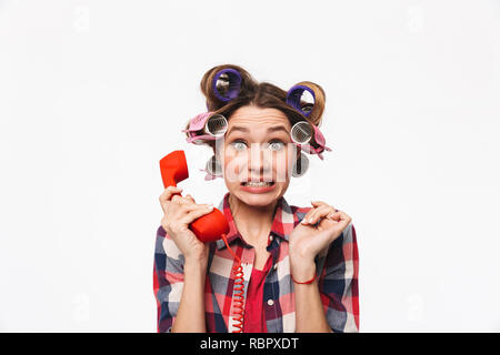 Disappointed housewife with curlers in hair standing isolated over white background, talking on a landline phone Stock Photo